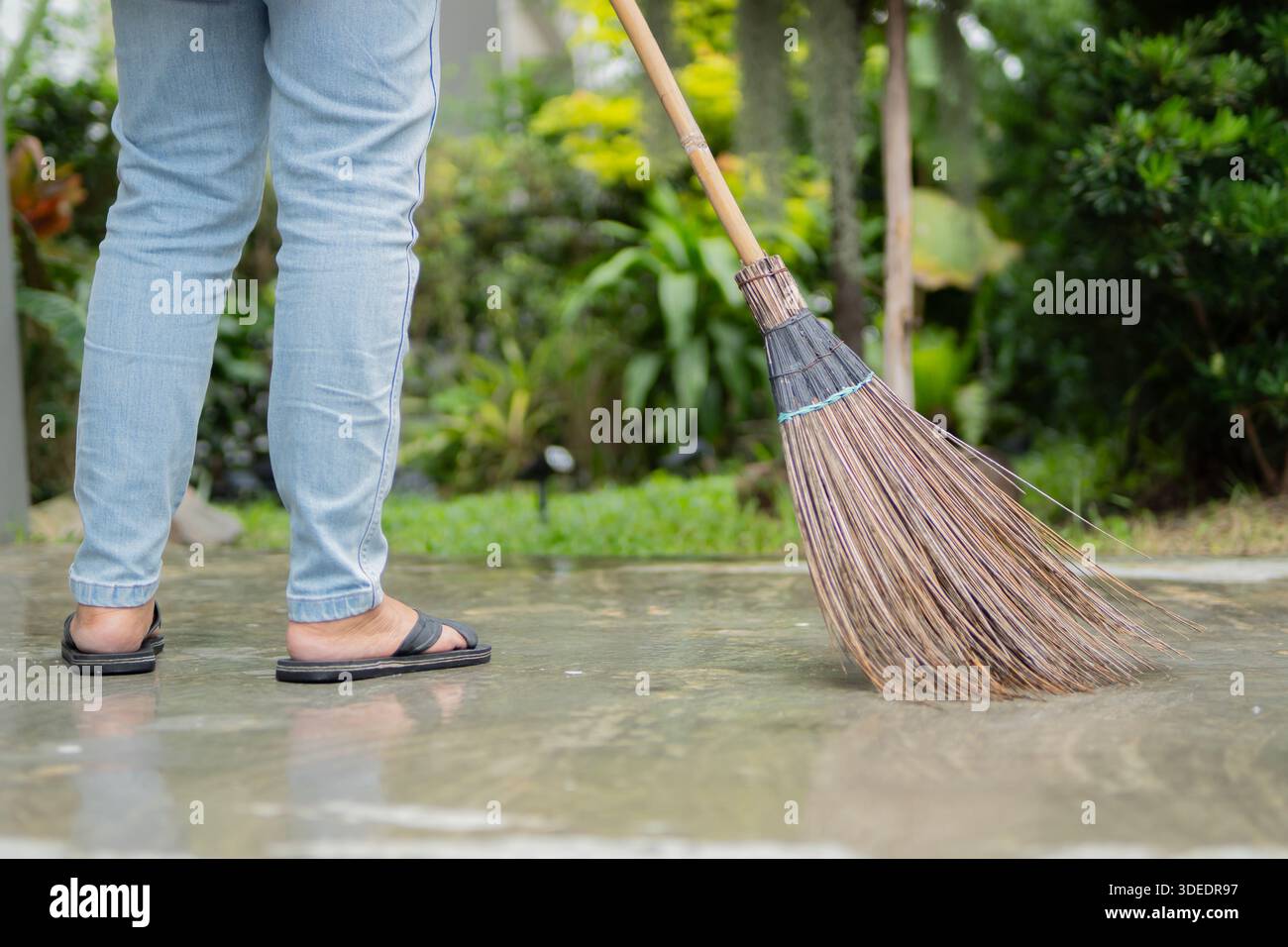 Vista laterale di una donna con scopa dura naturale durante il soffio su un sentiero del giardino. Una tradizionale scopa di paglia. Foto Stock