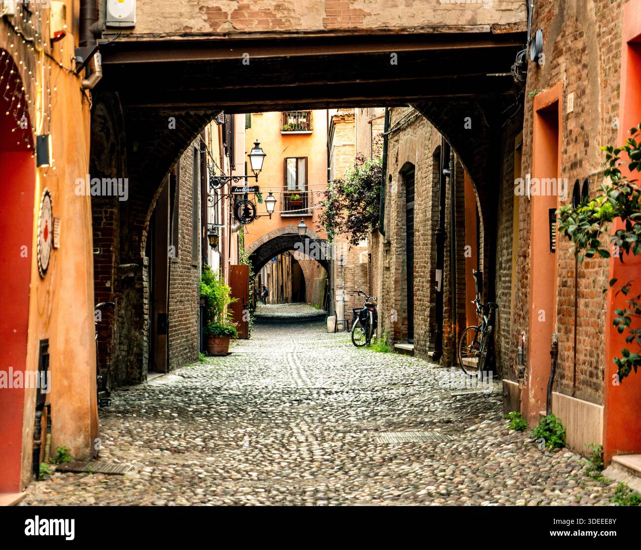 Una pittoresca strada acciottolata in una città vecchia, caratterizzata da passerelle ad arco, edifici in mattoni e vegetazione, con biciclette parcheggiate nelle vicinanze Foto Stock