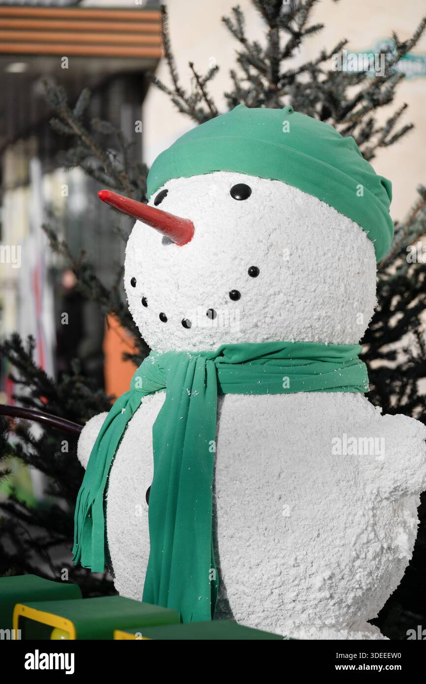 Scatto verticale di un pupazzo di neve decorativo che indossa un cappello verde e una sciarpa con un naso di carota rossa. Decorazioni natalizie e di Capodanno con sfocature Foto Stock