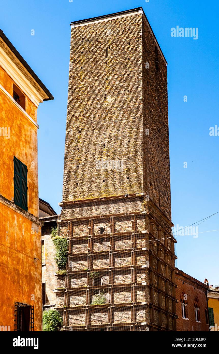 Un'alta, antica torre di pietra con una superficie strutturata, circondata da edifici colorati sotto un cielo blu chiaro Foto Stock