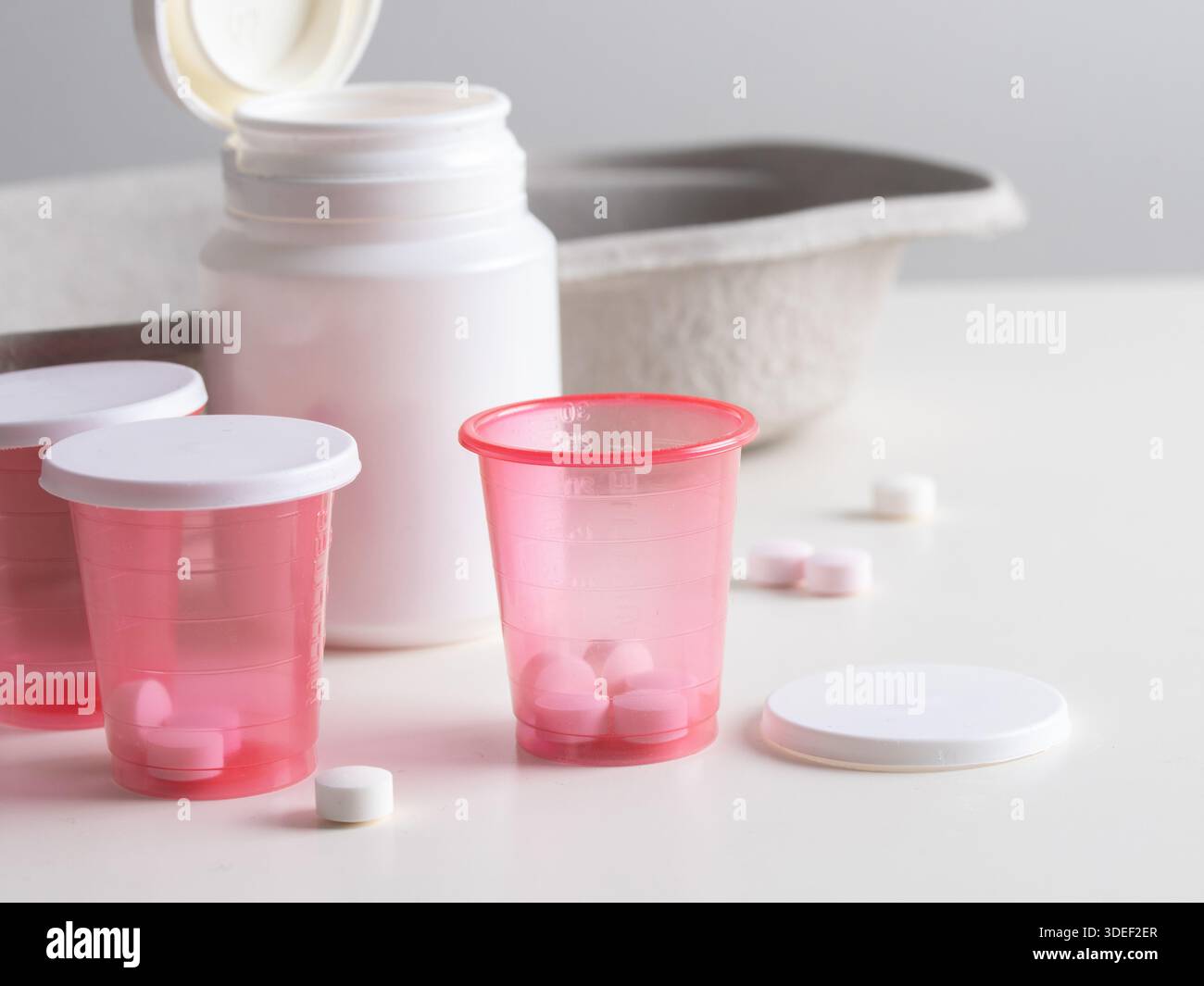 Bottiglia di plastica bianca con coppette medicinali rosse contenenti compresse bianche. Piccole pillole bianche sparse su una superficie bianca. Medicale o farmaceutico S Foto Stock
