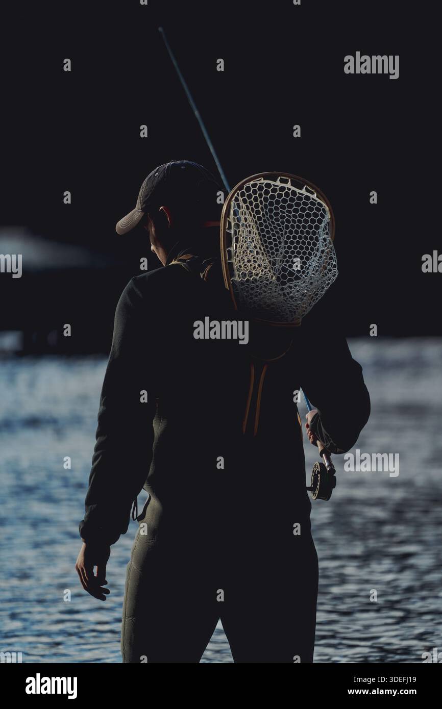 Un pescatore di mosca si trova in un fiume che regge una canna da pesca e una rete, sagomata contro l'acqua scura e la luce naturale. Foto Stock
