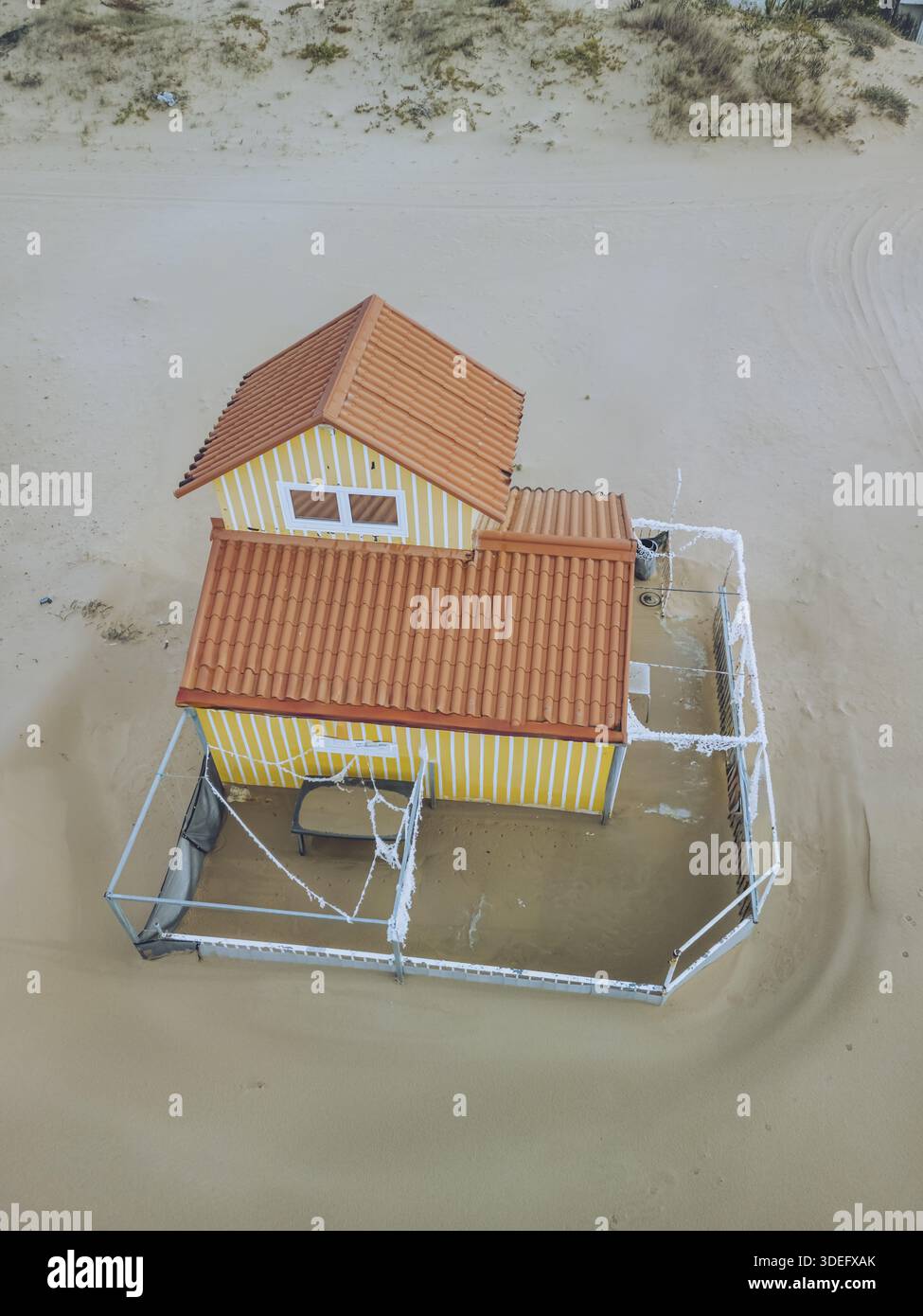 Vista aerea di una casa gialla in miniatura con un tetto arancione si trova all'interno di dune di sabbia, un solitario faro di presenza umana, Costa da Caparica, S. Foto Stock