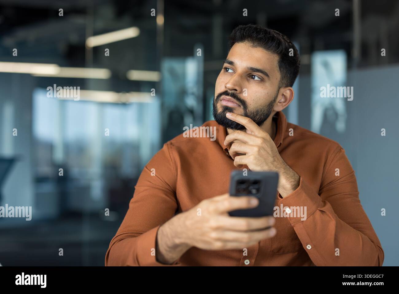 Giovane uomo d'affari adulto che tiene in mano uno smartphone mentre cerca di creare idee nuove o prende decisioni importanti in un moderno ufficio aziendale Foto Stock