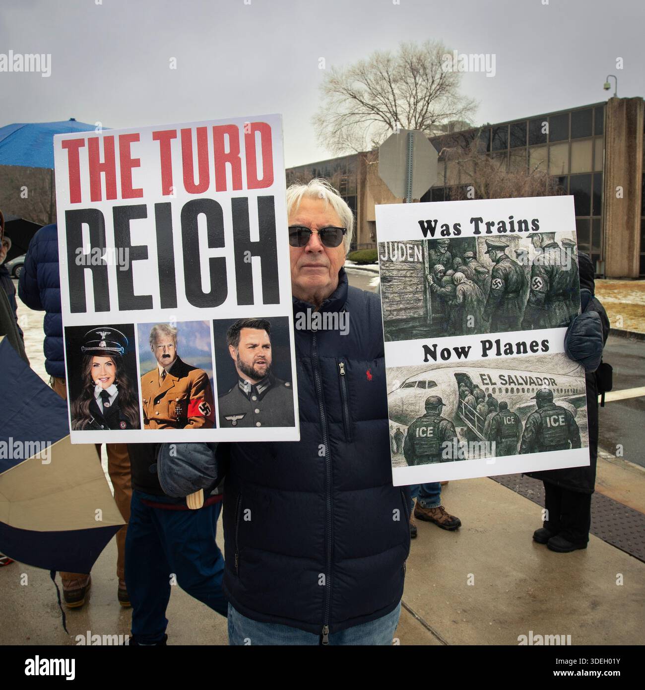 Burlington, Massachusetts, Stati Uniti. 7 GENNAIO 2026. 38a manifestazione settimanale ANTIGHIACCIO «testimone» in una fredda giornata di gennaio di fronte all'ufficio di immigrazione e applicazione delle dogane del New England, 26 km a nord-ovest di Boston, ma. A causa della pioggia gelida, la manifestazione settimanale è stata ufficialmente cancellata, ma fino a 75 persone si sono ancora riunite pacificamente fuori dalla struttura per due ore il mercoledì a mezzogiorno. Un manifestante in piedi all'ingresso della struttura ha un cartello con le foto di Kristi Noem, Donald Trump e JD Vance raffigurati come Nazis Credit: Chuck Nacke / Alamy Live News Foto Stock