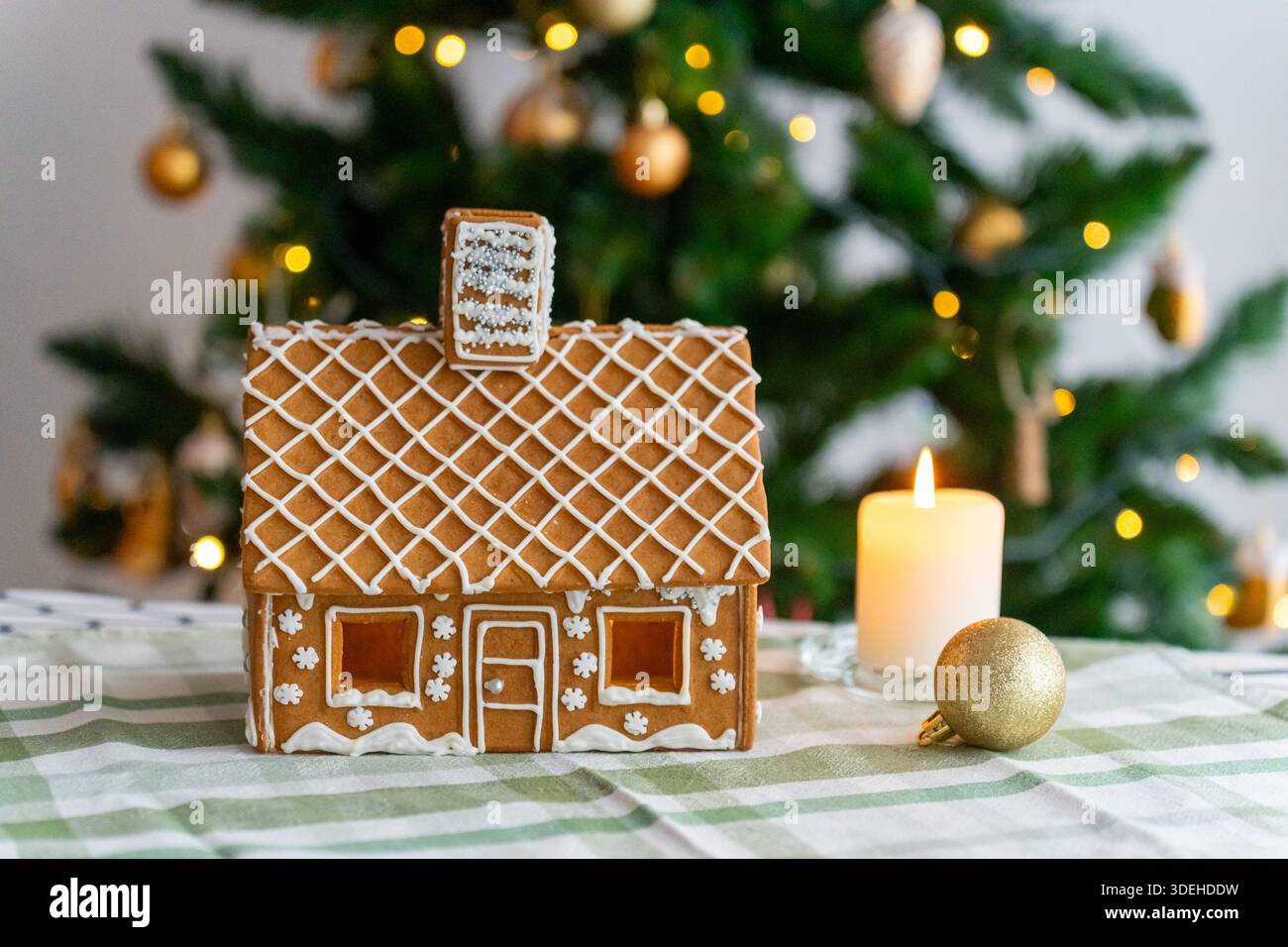 Casa di pan di zenzero fatta a mano con tetto a glassa con motivo a diamante e finestre luminose accanto a candela bianca e bauble oro su tovaglia verde a quadri con bok Foto Stock