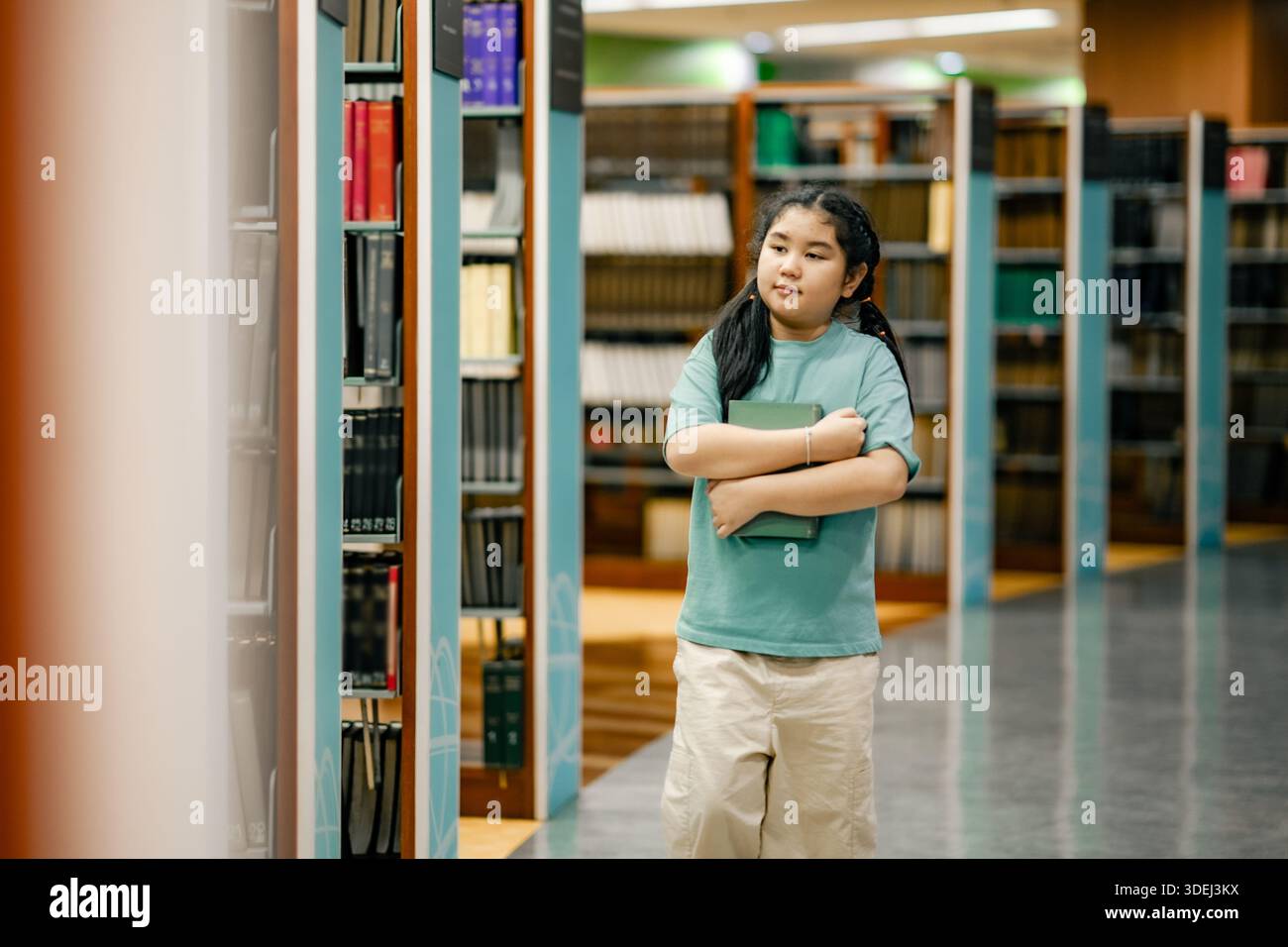 Studentessa asiatica che si muove attraverso gli scaffali della biblioteca con il libro in braccio. L'aspetto evidenzia l'indipendenza l'attenzione la curiosità l'istruzione la crescita la scuola positiva Foto Stock