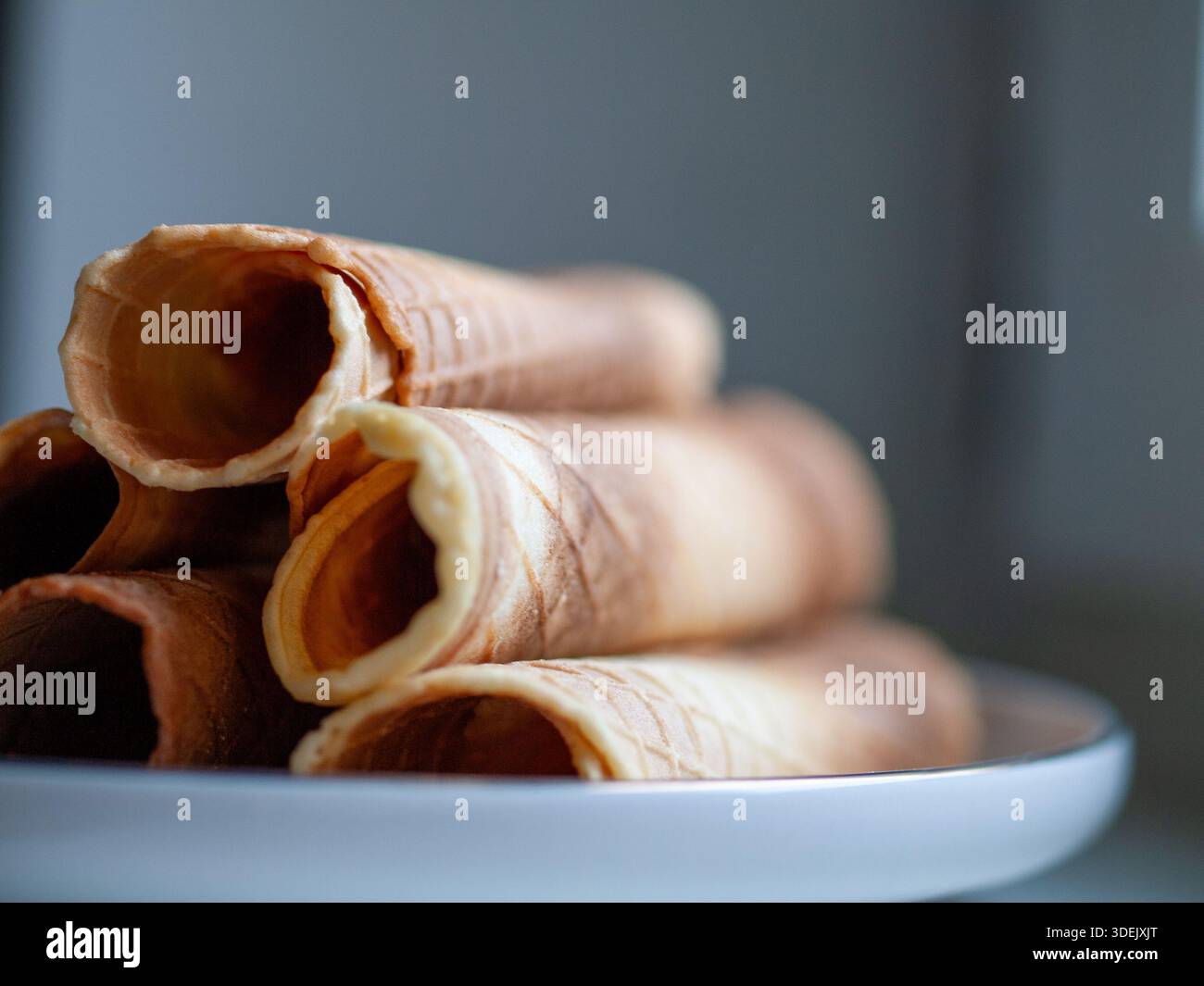 Coni arrotolati di wafer disposti su un piatto bianco su uno sfondo blu neutro, natura morta del cibo minimalista. Foto Stock