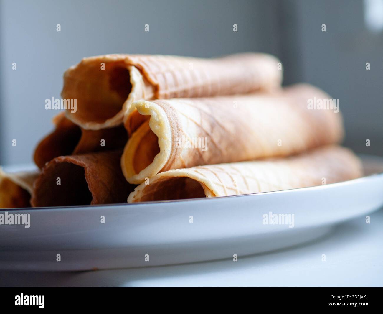 Coni di wafer arrotolati impilati su una piastra di ceramica bianca, natura morta di alimenti ravvicinata con profondità di campo ridotta. Foto Stock