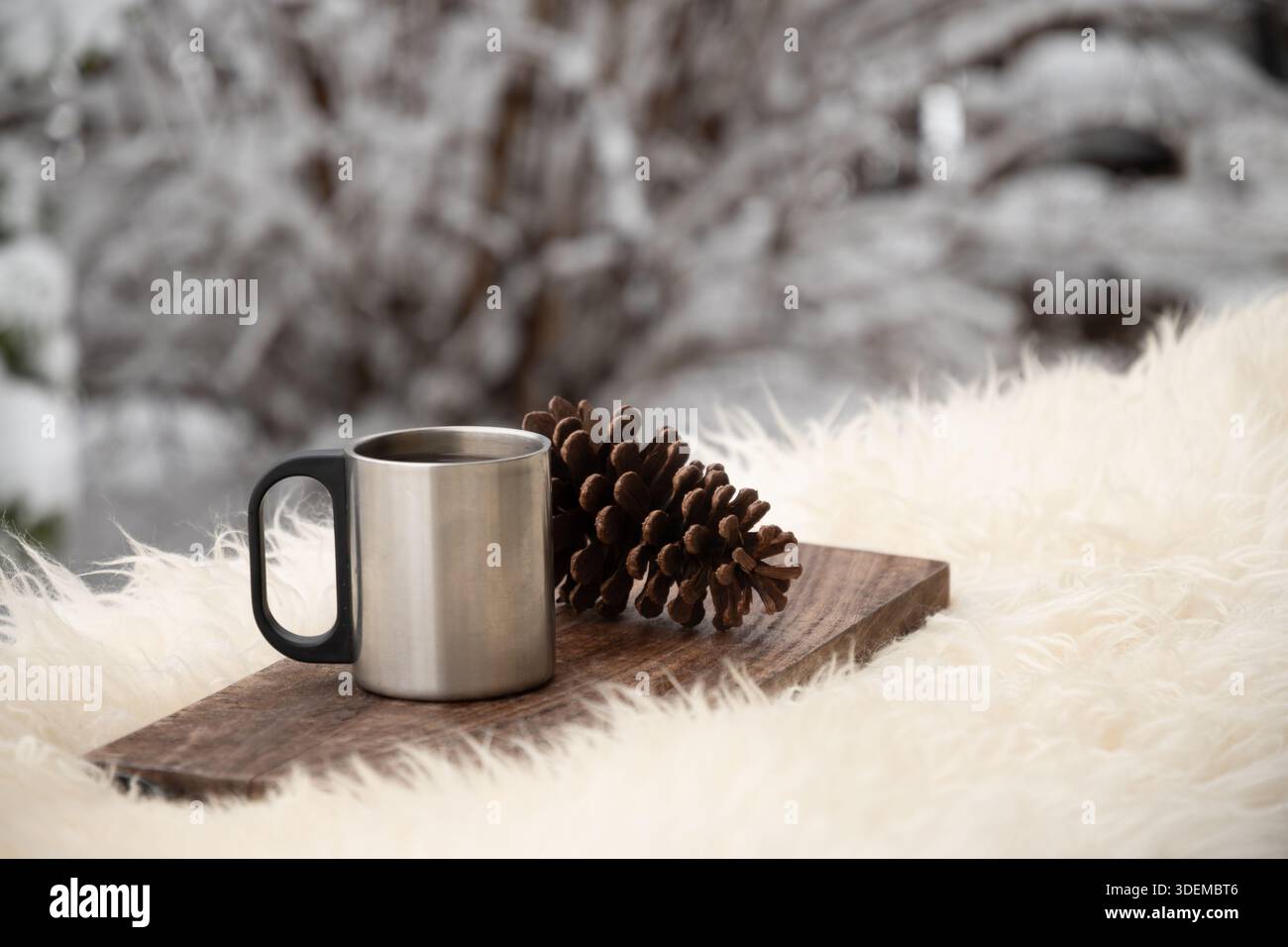 Tazza in metallo con bevanda calda e cono di pino su tavola di legno all'aperto. Composizione invernale minimalista con sfondo innevato e spazio di copia. Foto Stock