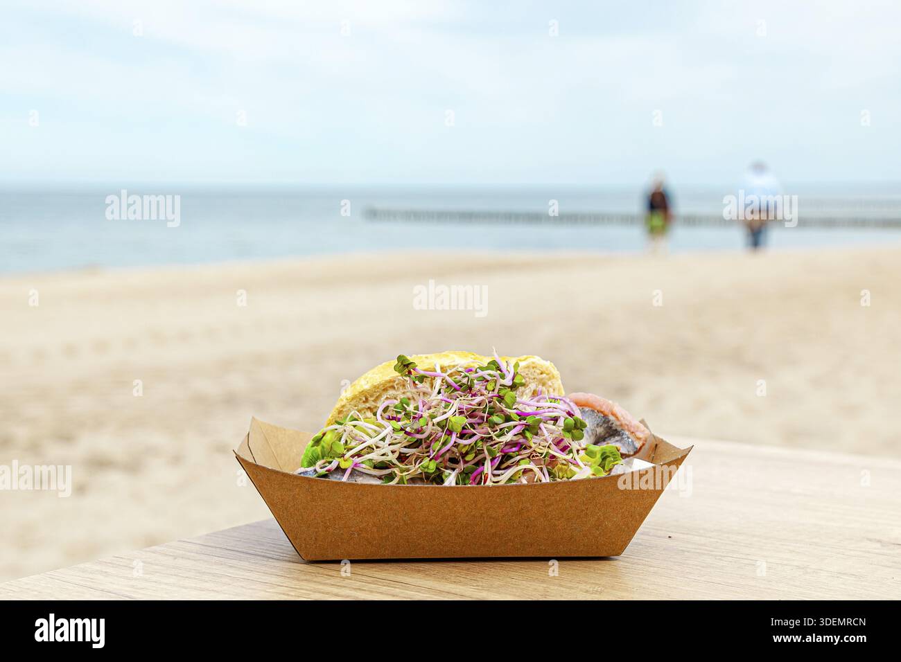 Involtino di pesce in una scatola per sandwich su un tavolo su una spiaggia. ripieni in scatole ecologiche di carta artigianali. Tradizionale cibo di strada della germania settentrionale. panino di grano con porticciolo Foto Stock