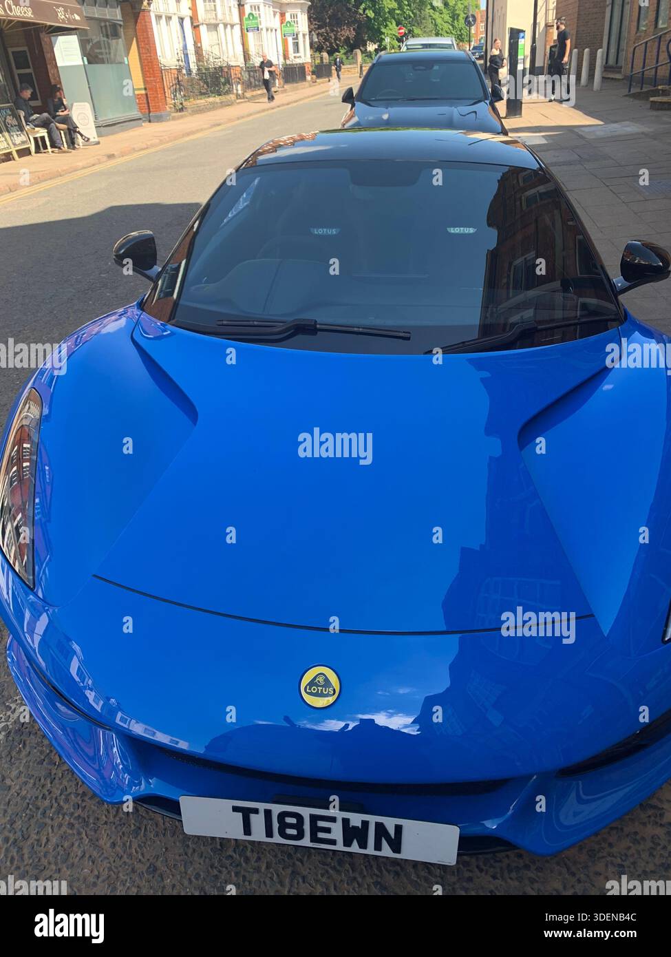 Una vivace auto sportiva blu con accenti neri e pinze dei freni gialle parcheggiata in una strada di fronte a una tradizionale auto classica in mattoni rossi - Immagine stock catturata con smartphone