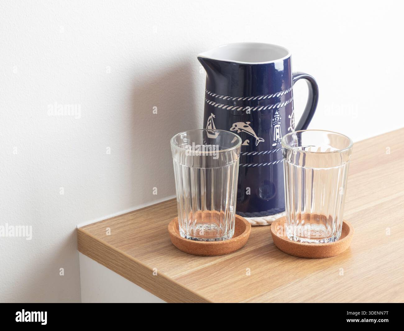 Una caraffa in ceramica a tema nautico è abbinata a due bicchieri da bere trasparenti e due sottobicchieri rotondi in sughero, per creare un elegante tavolo ispirato alla costa Foto Stock
