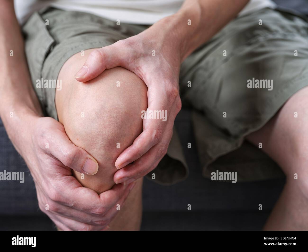 Un uomo di mezza età seduto su un divano, che tiene il ginocchio e la gamba con entrambe le mani a soffrire. Primo piano. Foto Stock