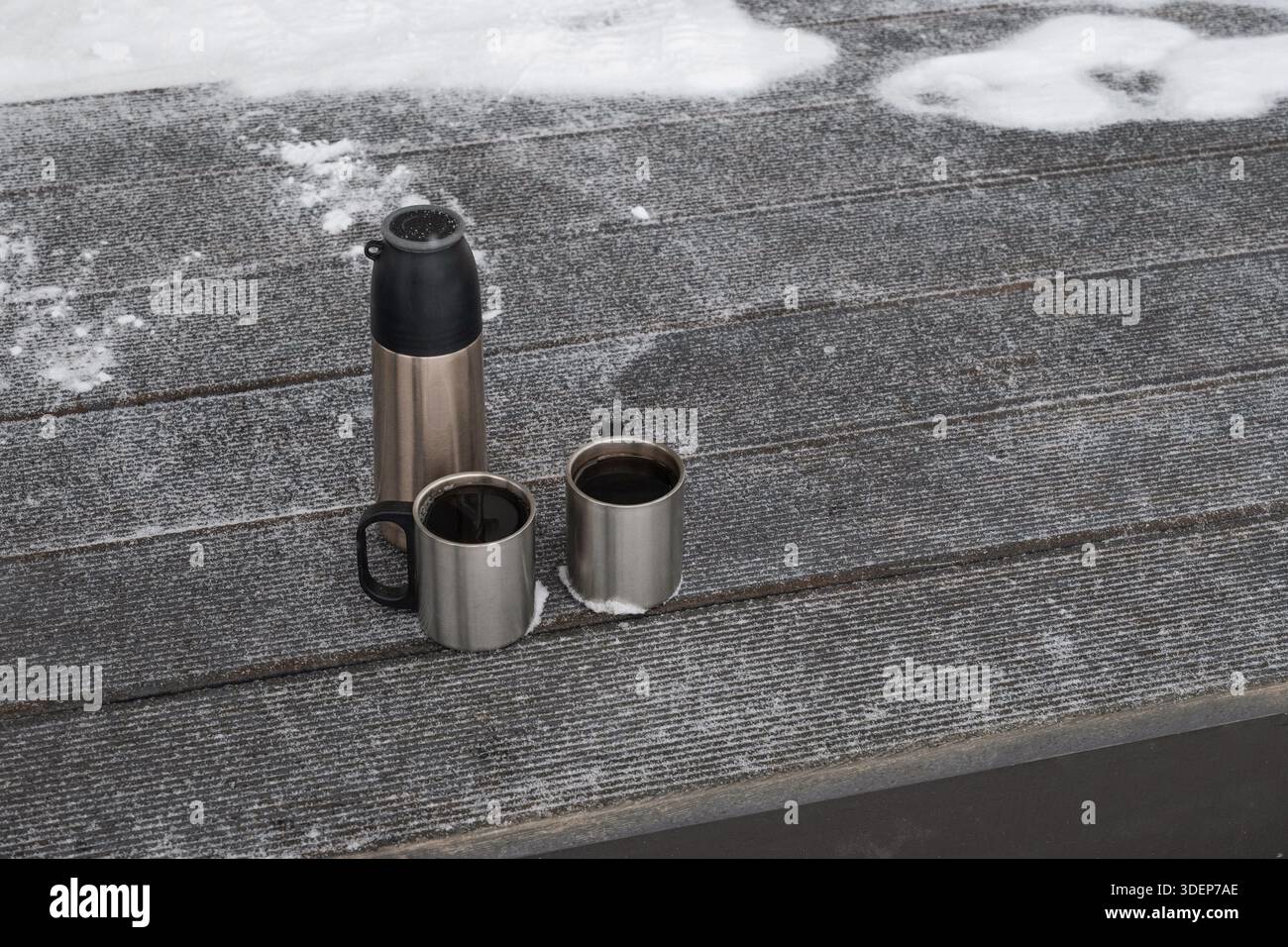 Thermos e tazze in metallo con bevanda calda collocate su una panchina di legno innevata all'aperto. Stile di vita invernale, clima freddo, relax all'aperto, composi minimali Foto Stock