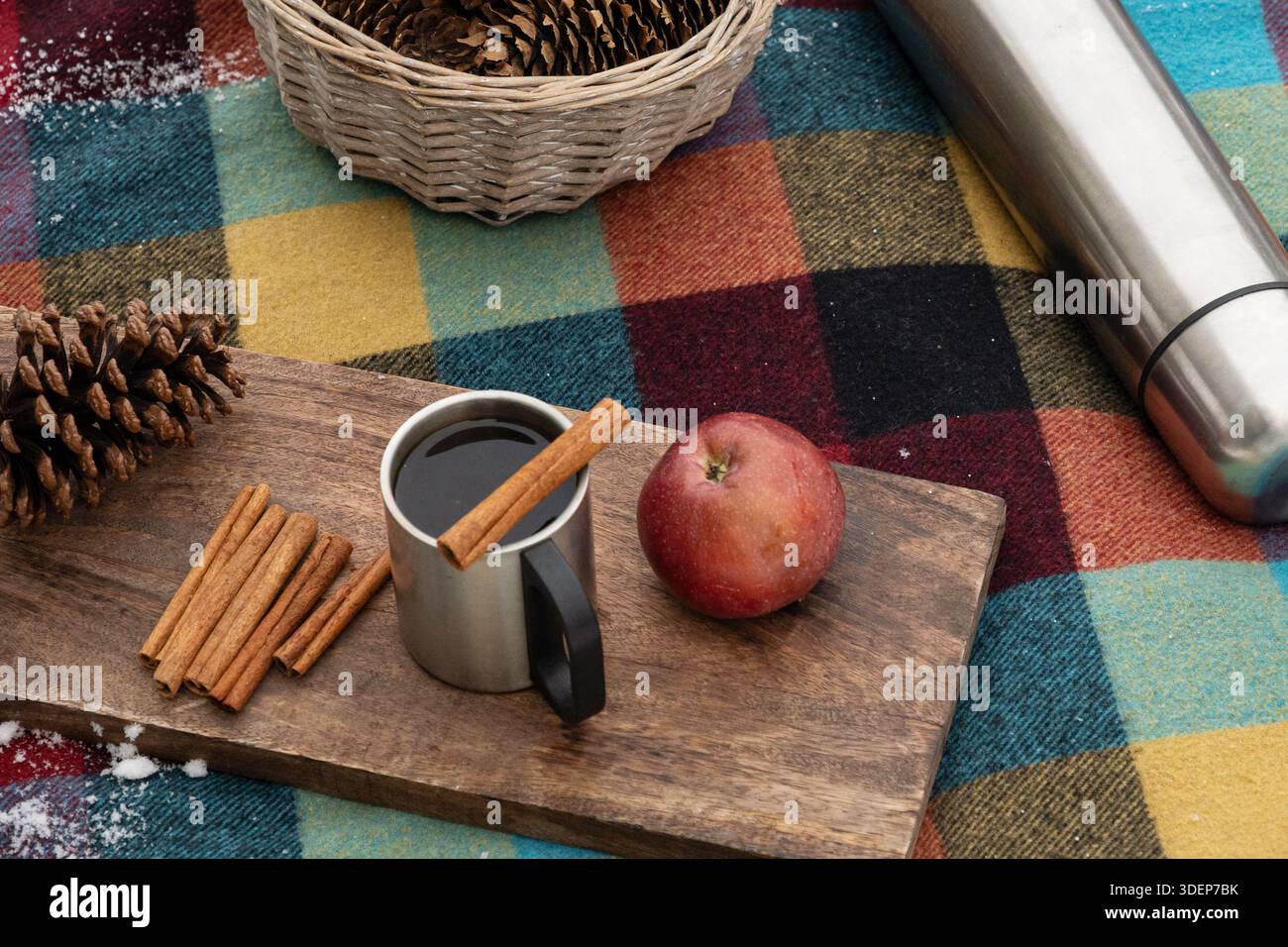 Tazza di metallo con bevanda invernale calda, bastoncini di cannella e mela su tavola di legno e coperta a quadri. Accogliente picnic invernale, ingredienti naturali, festa della stagione Foto Stock