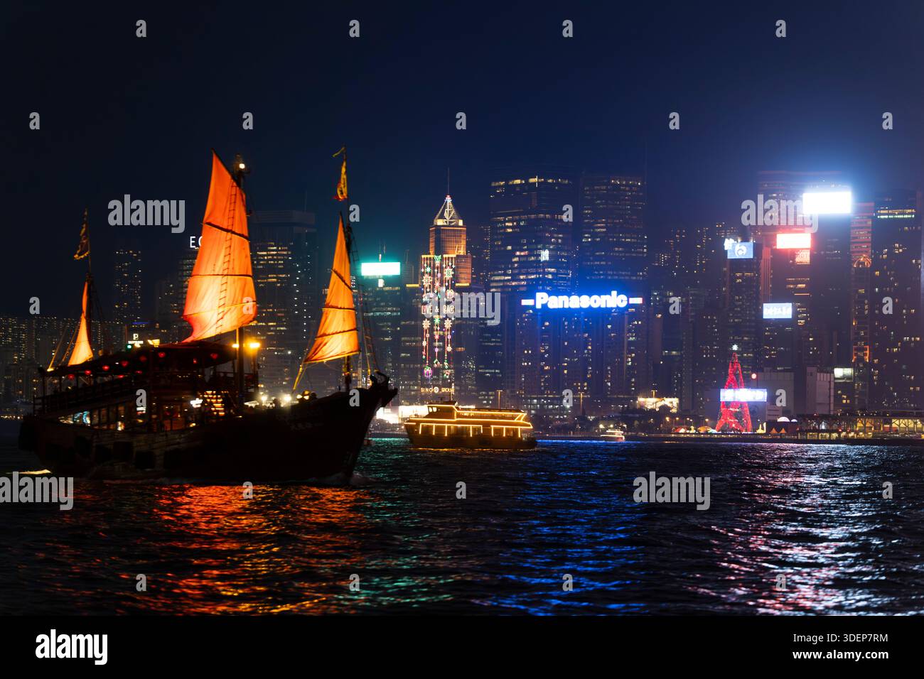 Una tradizionale barca spazzatura naviga nel Victoria Harbour, Hong Kong, con lo skyline illuminato di Tsim Sha Tsui sullo sfondo. Foto Stock