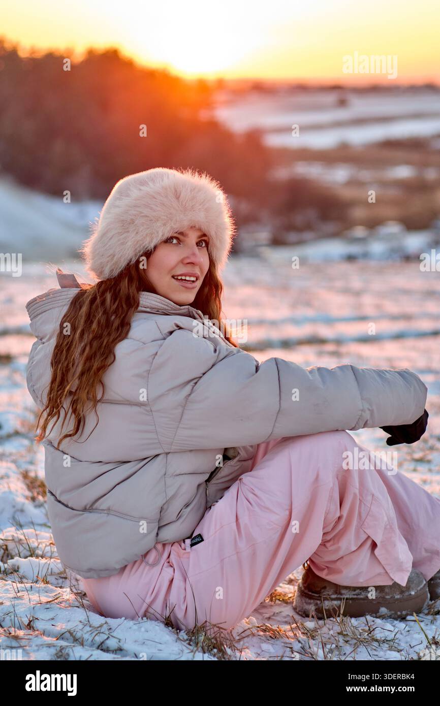 Una giovane donna è seduta sulla neve, vestita con un caldo abbigliamento invernale che include un cappello di pelliccia e un'accogliente giacca a piumino. Si sta godendo un inverno pittoresco Foto Stock