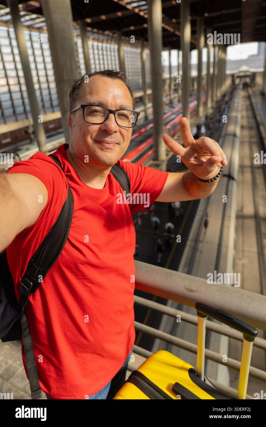 Un uomo di mezza età che indossa gli occhiali da lettura sorride con calma mentre scatta un selfie e fa un segno di pace in una moderna stazione ferroviaria. Viaggi rilassati e urba Foto Stock