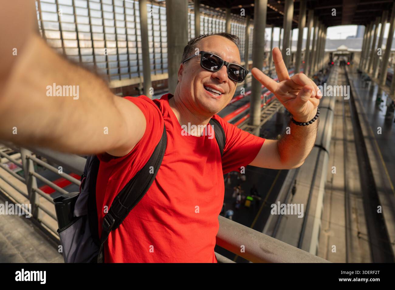 Un uomo di mezza età che indossa gli occhiali da sole si fa un allegro selfie in una stazione ferroviaria trafficata. Viaggi moderni, stile di vita urbano, viaggi estivi e vacanza Foto Stock
