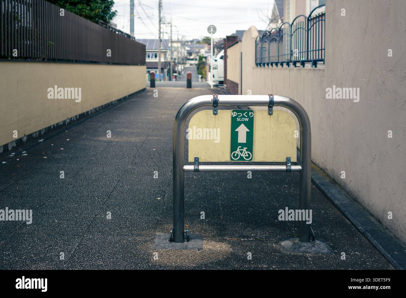 Un cartello che ricorda ai ciclisti di andare piano. La scena è un vicolo a Yokohama, circondato da mura e edifici beige sotto un cielo nuvoloso. Foto Stock