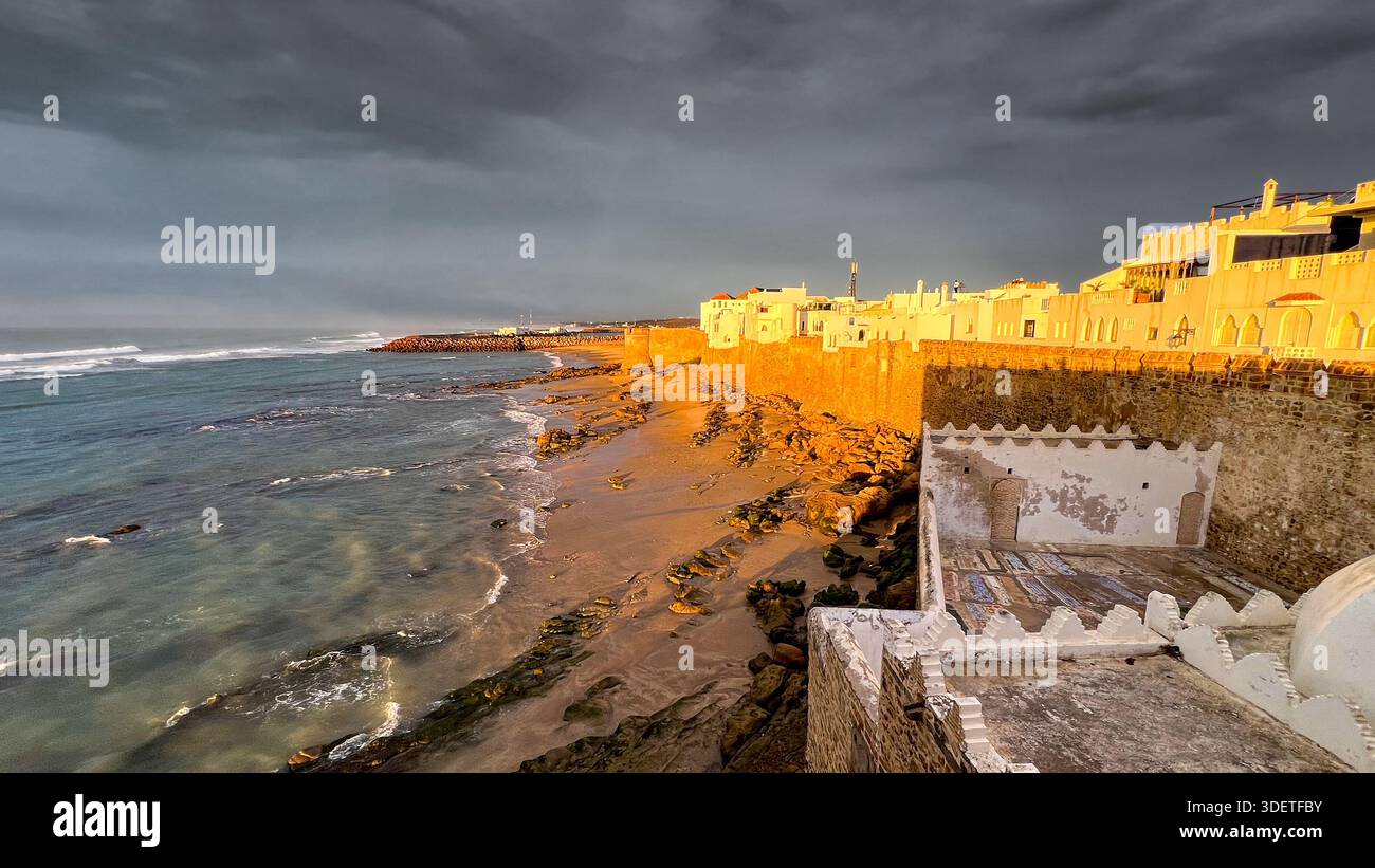 I bastioni della città fortificata di Asilah sono illuminati dal sole che tramonta mentre le nuvole di pioggia si allontanano. Foto Stock