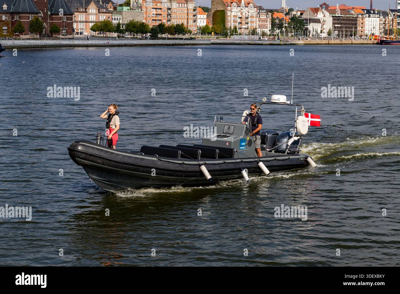 Aarhus, Danimarca - 15 agosto 2025: Piccolo motoscafo che trasporta due persone con bandiera danese, crociera vicino al porto cittadino, edifici residenziali sullo sfondo. Foto Stock