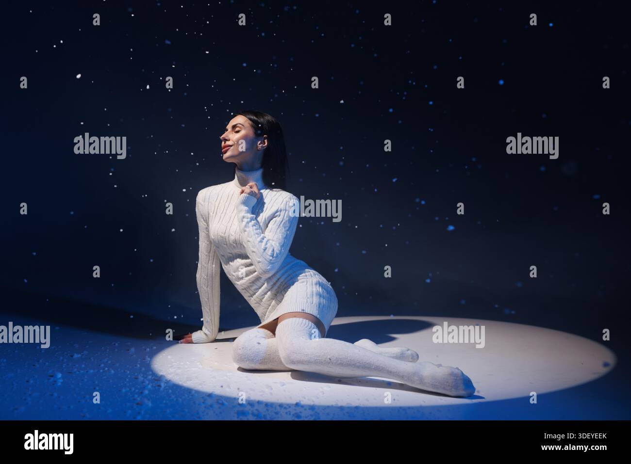 Donna con maglione bianco e calze al ginocchio che si siede contro la neve volante, concetto invernale freddo, Foto Stock