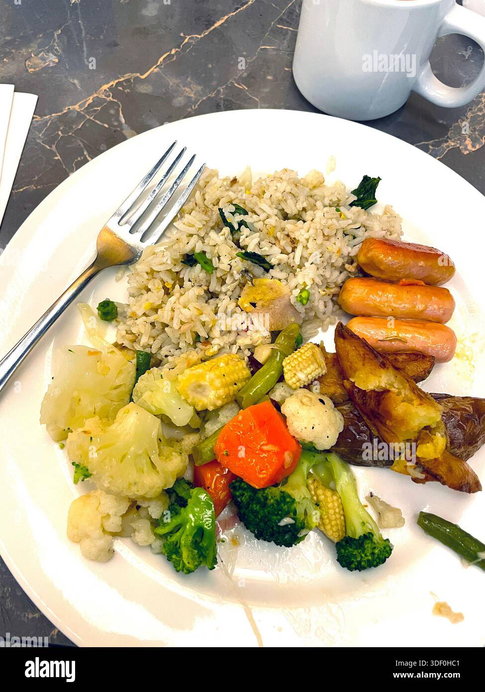 Piatto di cibi misti con riso fritto, patate arrosto, mini salsicce e verdure assortite al vapore su un tavolo da pranzo Foto Stock