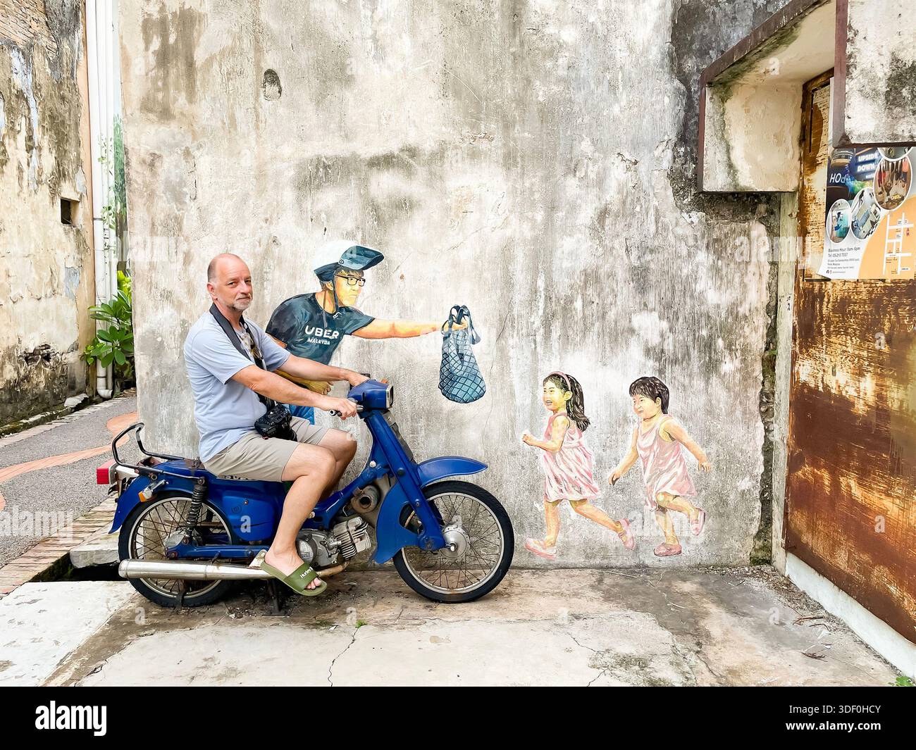Turista maschile su una moto d'epoca blu che posa di fronte al murale interattivo con i bambini e consegna cibo concubine Lane, Ipoh malesia Foto Stock