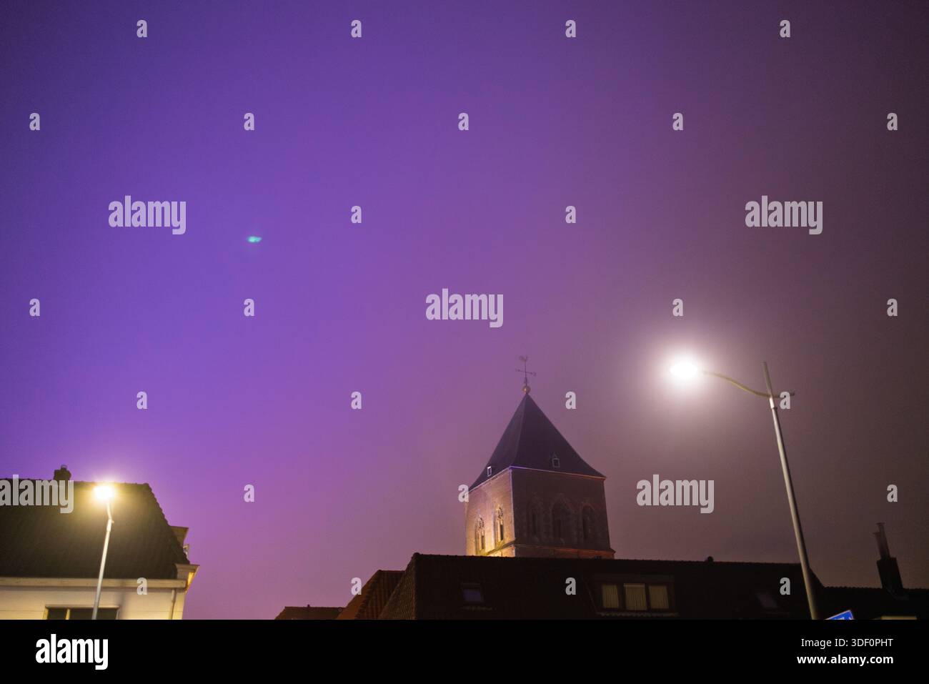 La torre della chiesa cittadina olandese illuminata di notte sotto il vivace cielo viola, i lampioni luminosi e la misteriosa luce blu-verde evocano intrighi d'atmosfera. Foto Stock