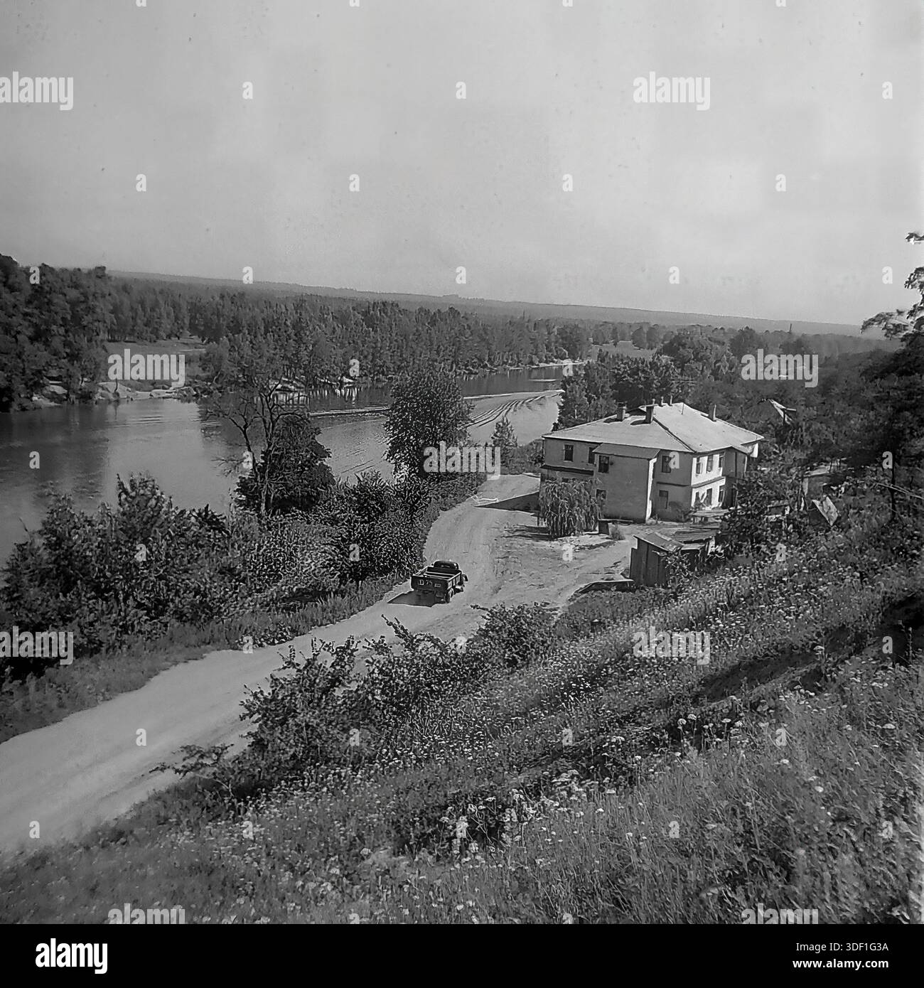 Sviatohirsk, regione di Donetsk, Ucraina, URSS. 1970 anni Vista panoramica dall'alto angolo della valle del fiume Seversky Donets. Un camion sovietico scuro guida lungo una tortuosa strada sterrata verso un edificio a due piani sulla riva del fiume. Un motoscafo si muove lungo il fiume sullo sfondo, lasciando una lunga scia bianca. La fitta foresta copre la sponda opposta. I pendii erbosi occupano del primo piano. Foto storica della vita della regione. Atmosfera autentica di lavoro estivo, svago e vita tranquilla nel Donbas prima della guerra. Foto Stock