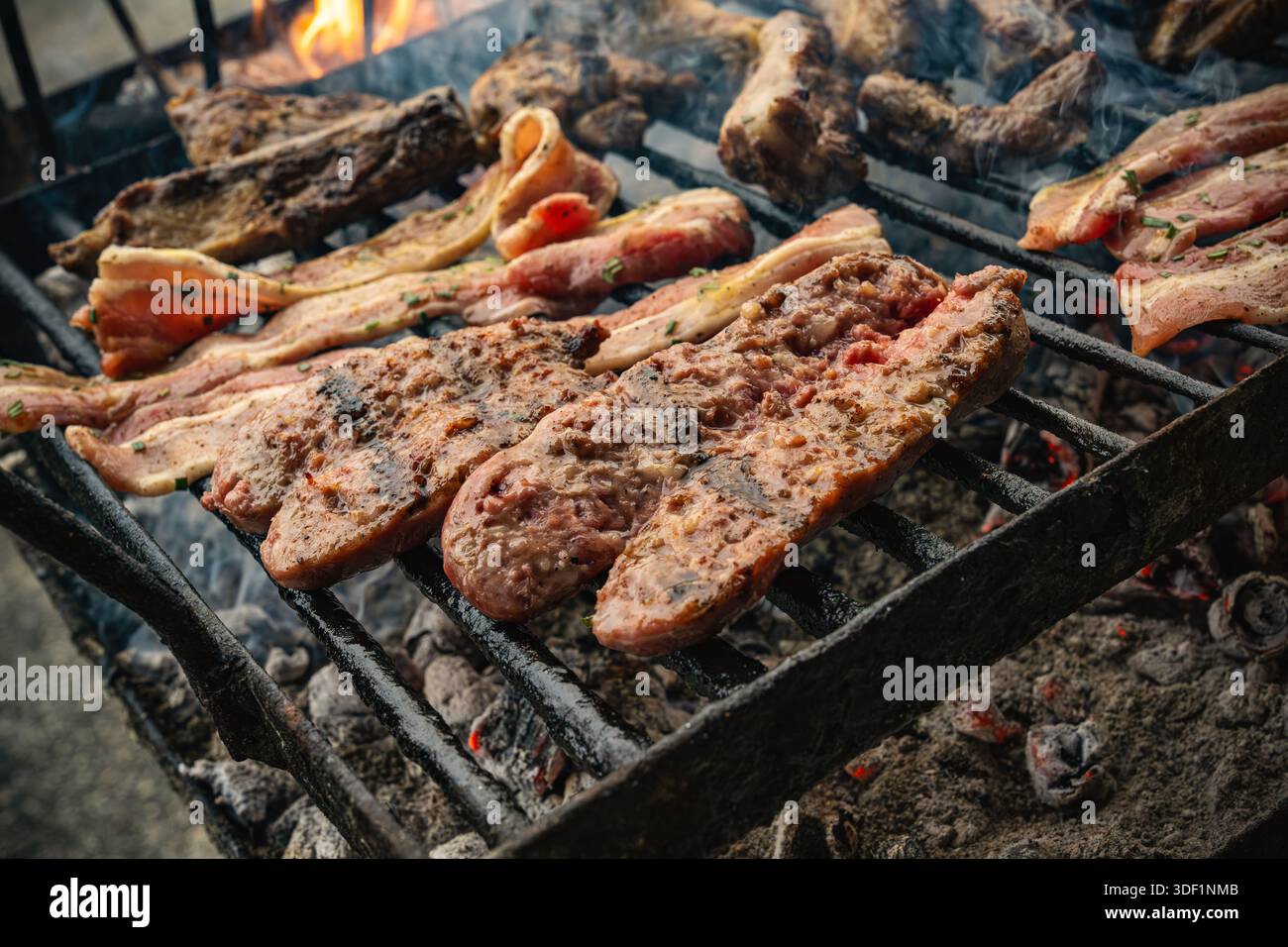 Carni sfrigolanti su una griglia a carbone con brace e fiamme accese, fumo che si arriccia mentre maiale e manzo cucinano all'aperto per una cucina calda e rustica estiva Foto Stock