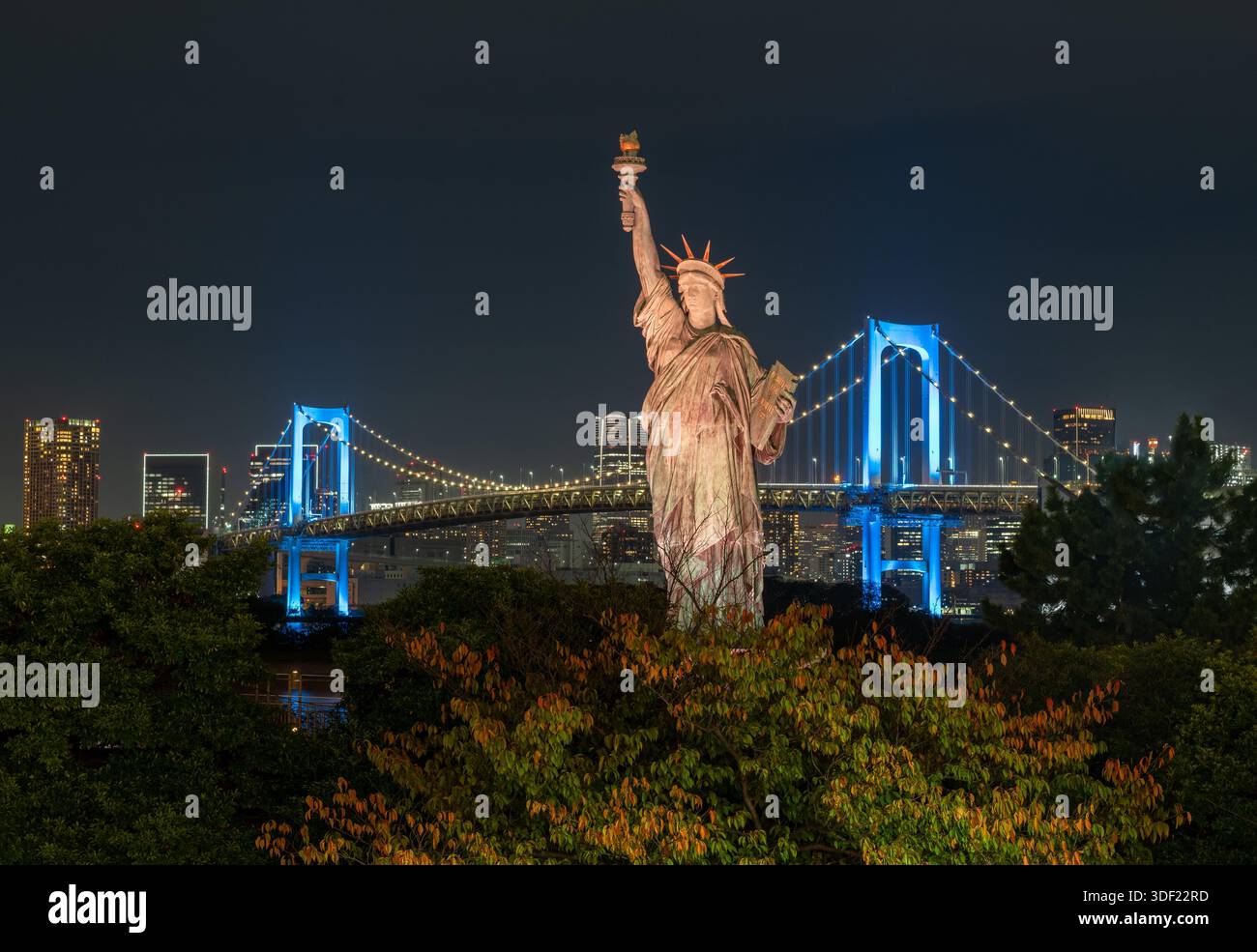 Vista della replica della Statua della libertà, inondata di luce soffusa, in mezzo a una lussureggiante vegetazione con il Ponte dell'Arcobaleno illuminato in blu, Tokyo, Tokyo, Foto Stock