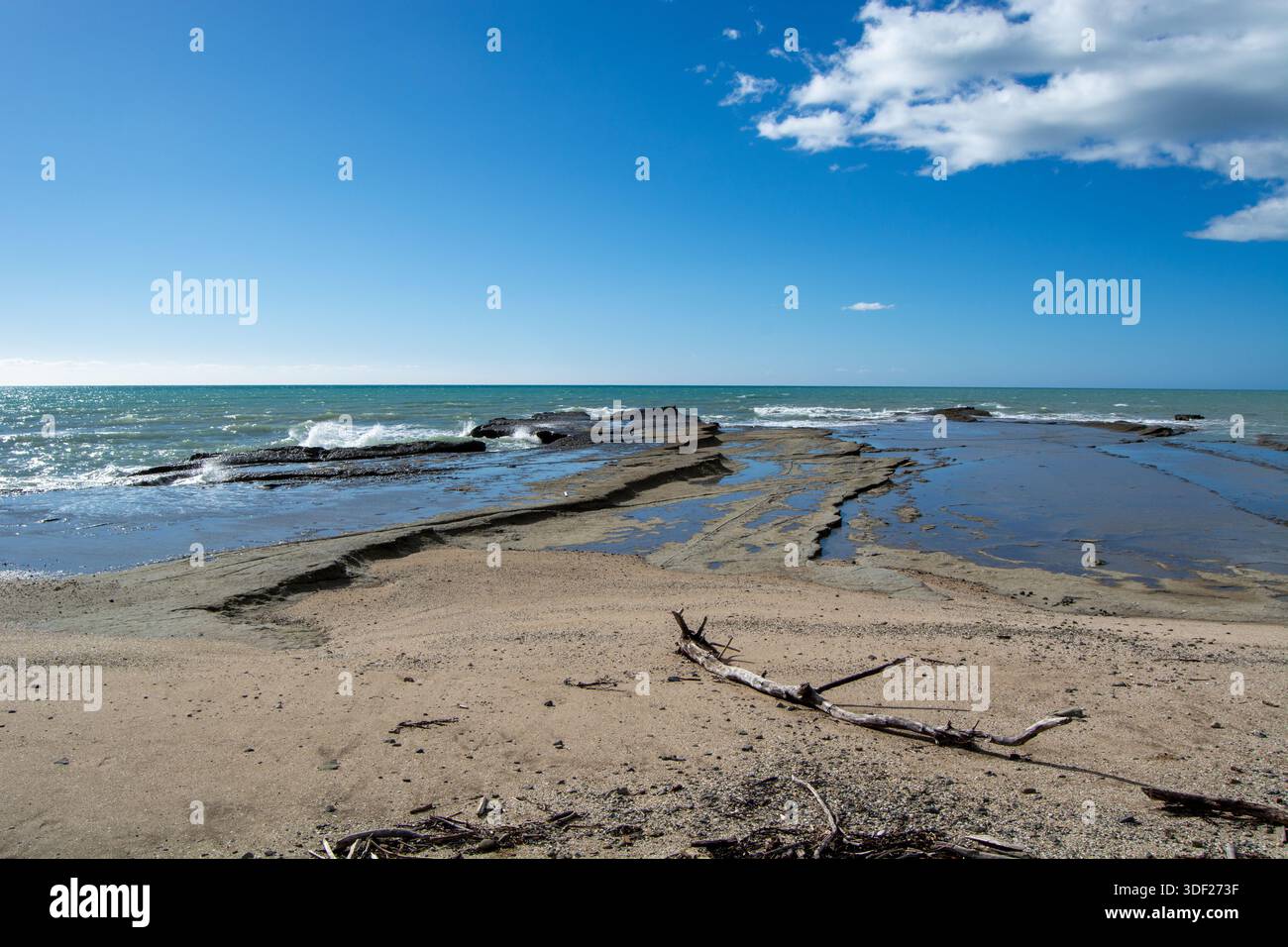 Un tranquillo paesaggio costiero caratterizzato da una costa rocciosa con piscine di marea, spiaggia sabbiosa e cieli azzurri. Onde dolci si infrangono contro le rocce sotto B. Foto Stock