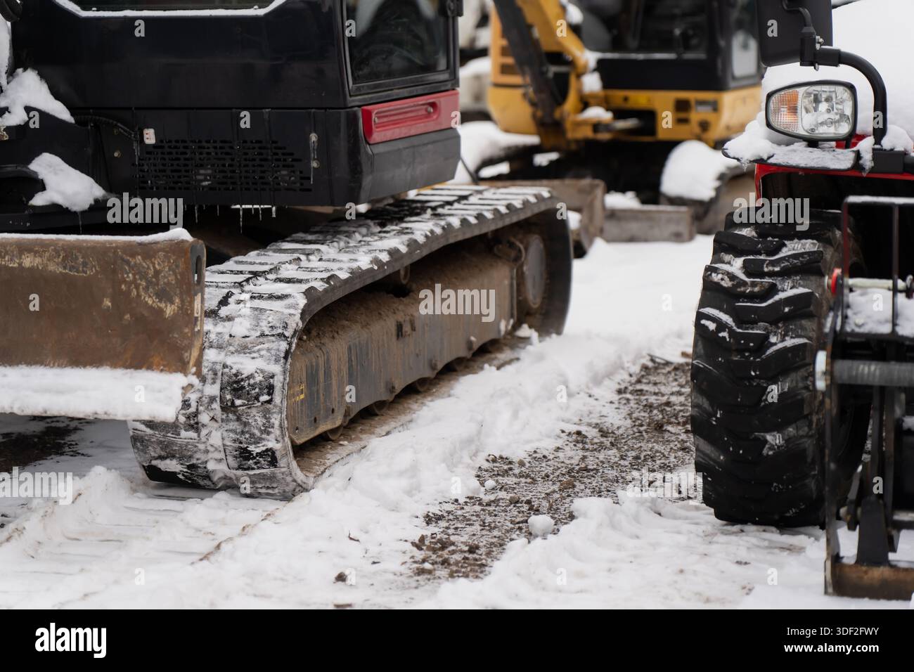 Dettagli sulle attrezzature da cantiere che mostrano i cingoli in acciaio dell'escavatore e le ruote compatte del veicolo su superfici innevate nei cantieri all'aperto nella stagione fredda Foto Stock