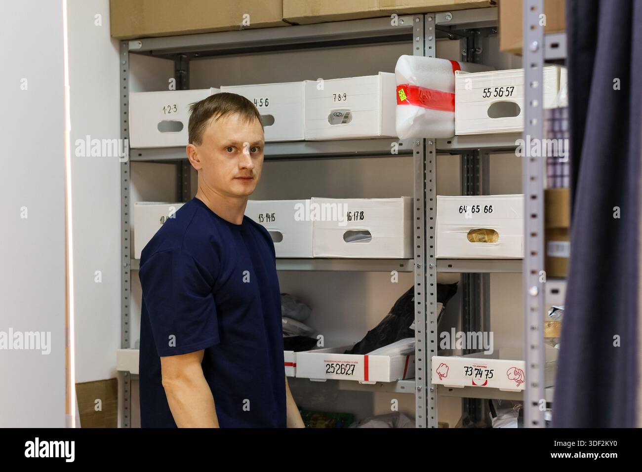 L'uomo in camicia blu posa neutralmente da scaffali metallici riempiti di scatole bianche e oggetti vari. Foto Stock