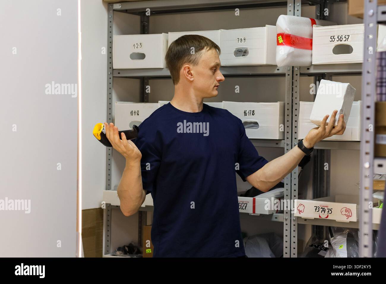 Il lavoratore in camicia navy mostra un piccolo oggetto bianco in una mano e un dispositivo portatile nell'altra nell'area rack. Foto Stock