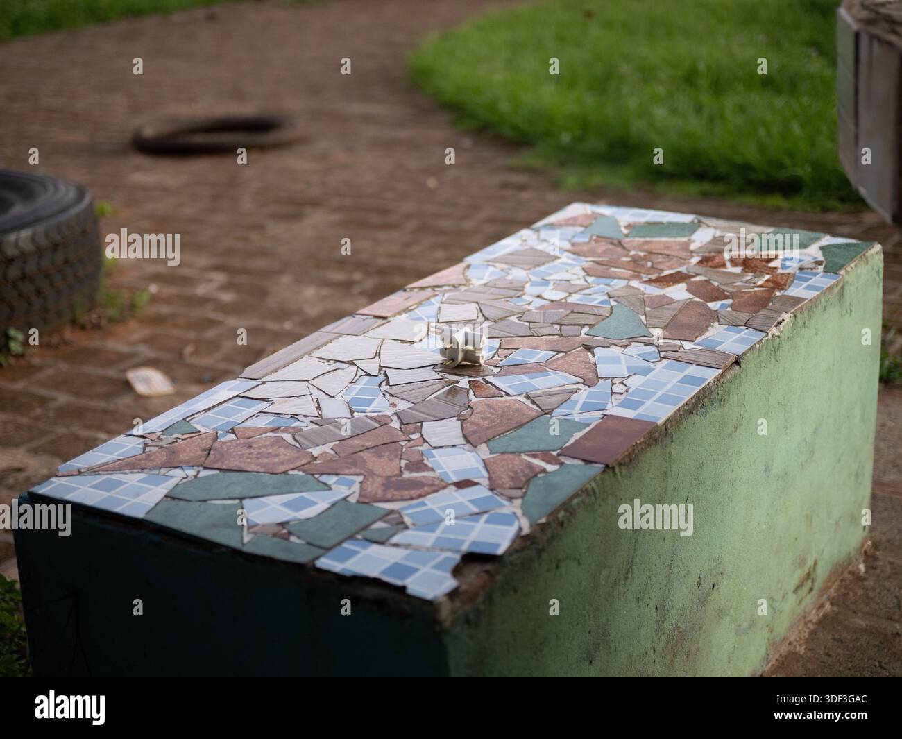 Panca del parco a mosaico con un piccolo oggetto metallico sulla superficie, che suggerisce un oggetto trovato in uno spazio pubblico all'aperto. Foto Stock
