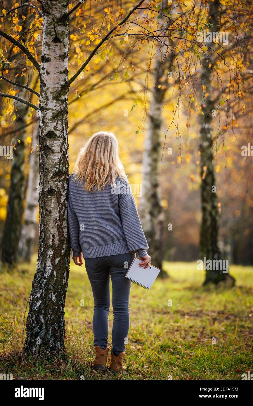 Donna in maglione in piedi nella foresta di betulle autunnale con un blocco note. Concetto di relax e di consapevole connessione con la natura Foto Stock