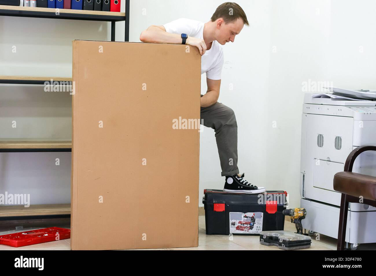 Uomo casual in equilibrio sulla cassetta degli attrezzi accanto alla cassetta di cartone verticale in ufficio. Giovane adulto con T-shirt bianca si staglia su una cassetta degli attrezzi nera, tenendo la parte superiore di grande T-shirt marrone Foto Stock