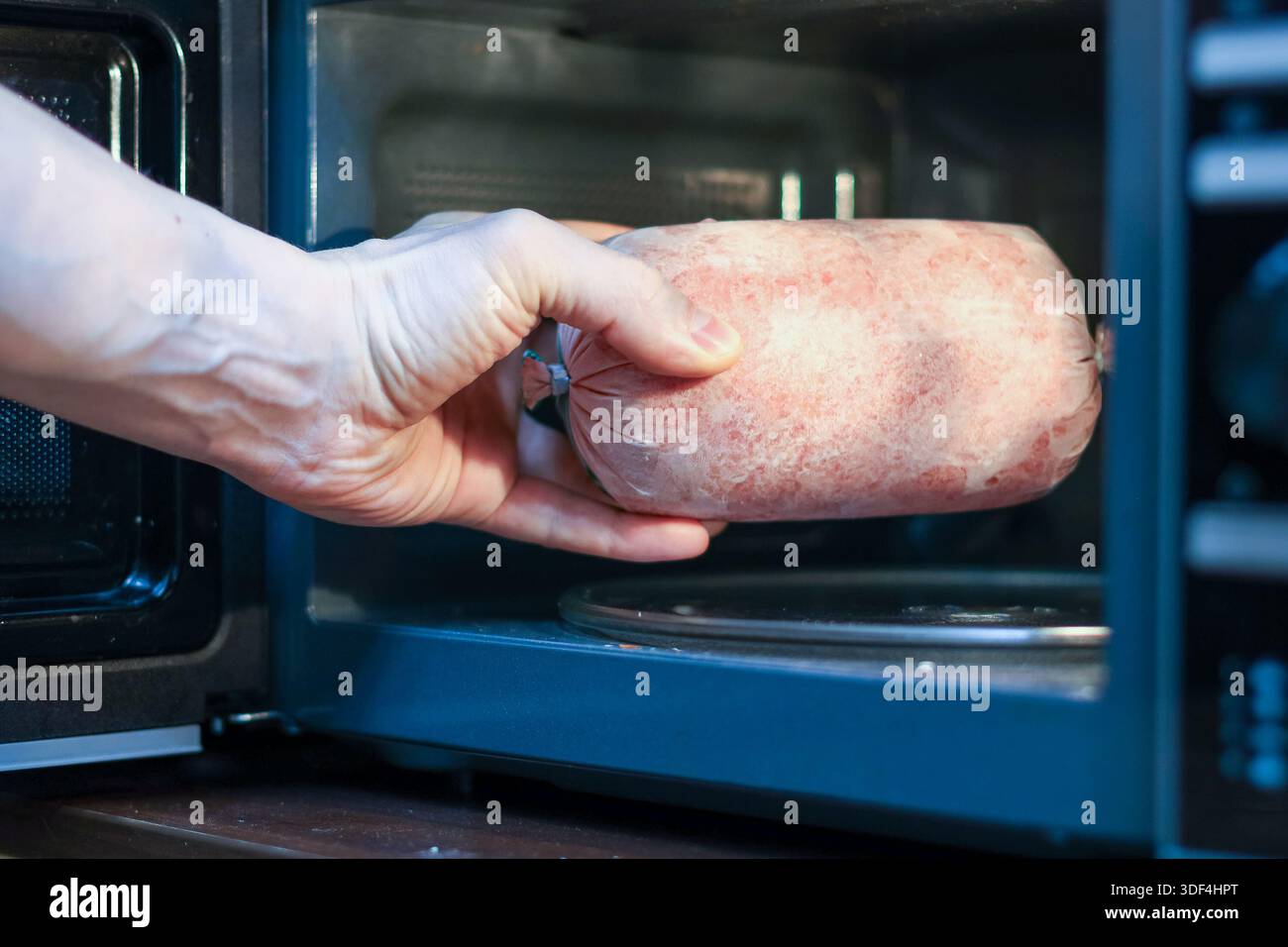 Inserire a mano una salsiccia rosa smerigliata nella porta aperta di un microonde su una superficie di legno. Foto Stock
