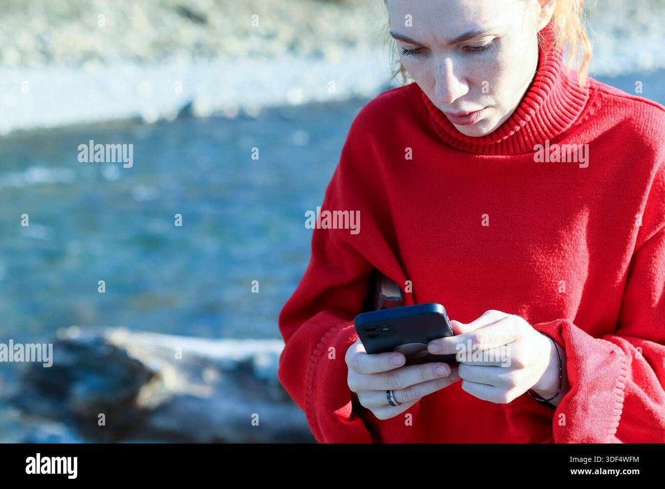 La giovane donna premurosa in un comodo maglione rosso controlla i messaggi sul suo smartphone mentre si trova accanto a una tranquilla riva del fiume con il fogliame autunnale sullo sfondo. Foto Stock