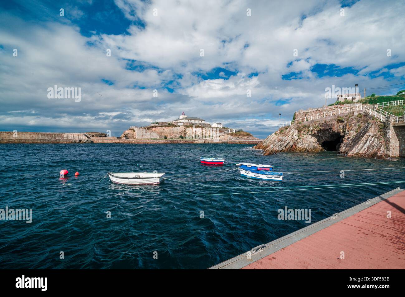 Il porto di Tapia de Casariego sul Mar Cantabrico presenta piccole barche da pesca ormeggiate in acque blu profonde, con il faro della città che si affaccia sulla roc Foto Stock