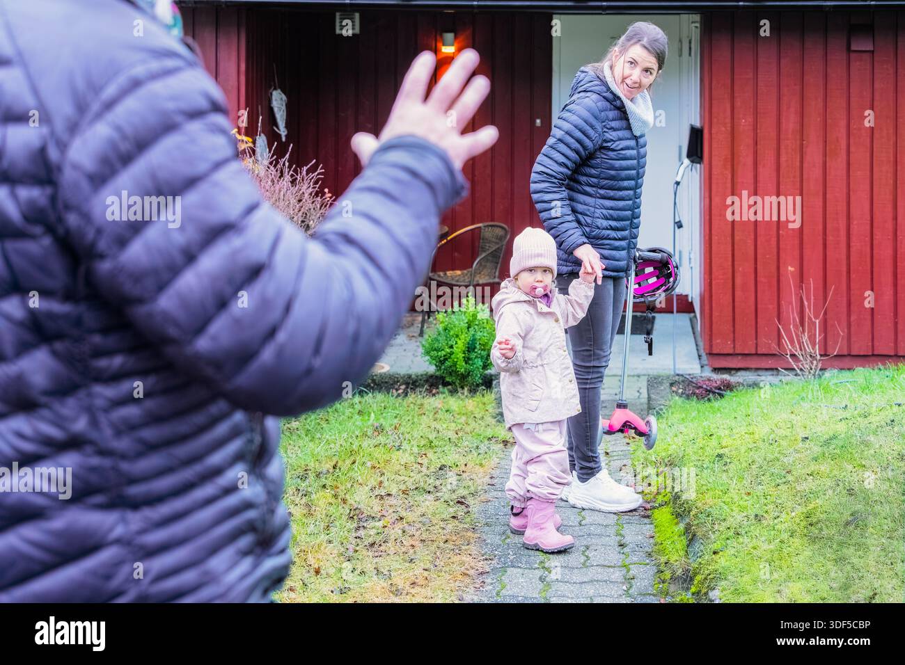 Una donna e un bambino camminano lungo un sentiero, salutando un uomo, con una casa affascinante e una vegetazione lussureggiante che creano una calda atmosfera familiare Foto Stock