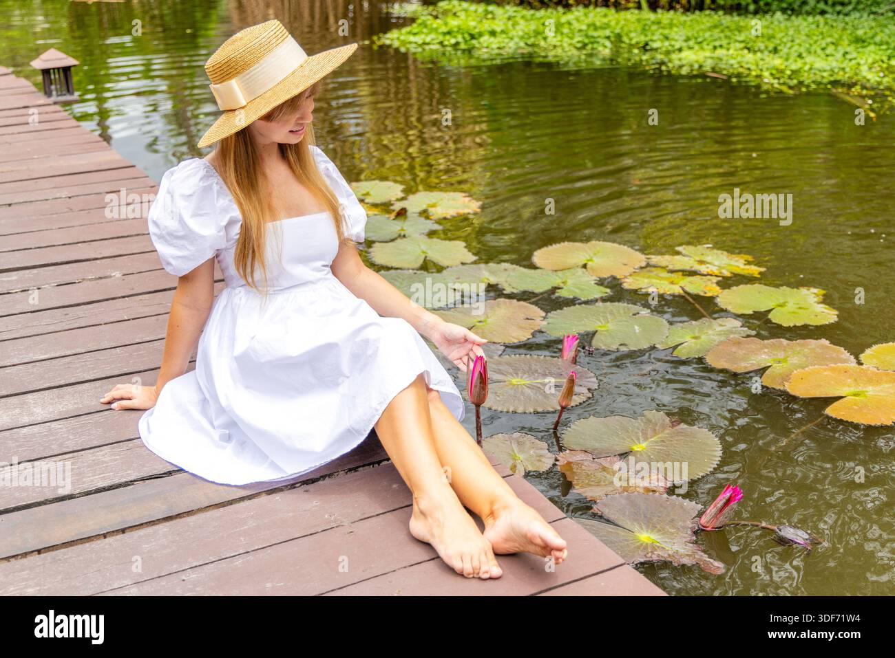 Donna in abito bianco seduta su un ponte di legno accanto a un laghetto con ninfee, in un ambiente naturale, con cappello di paglia, rilassante durante la giornata di sole. Foto Stock