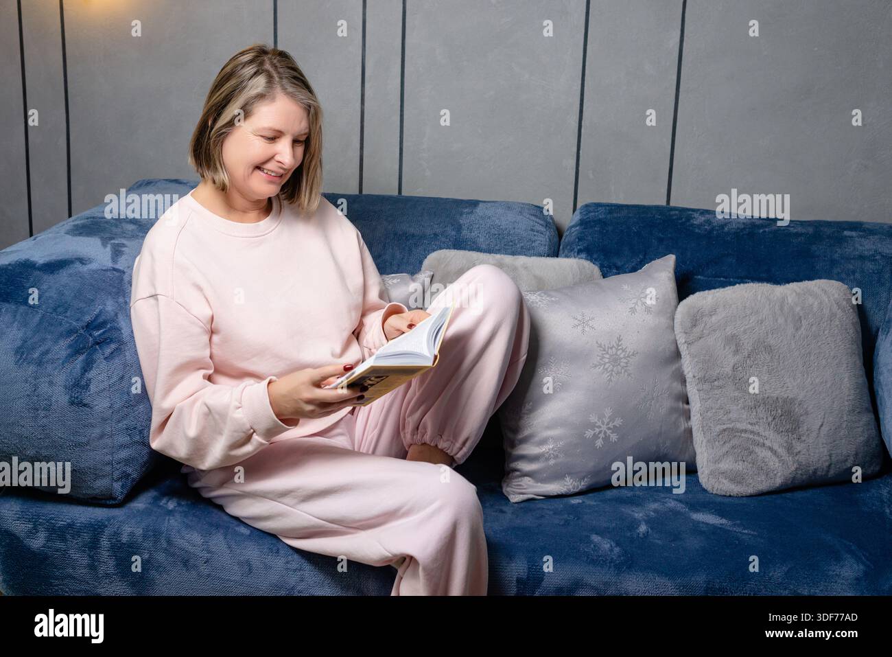 Donna che si rilassa sul divano mentre legge a casa, tranquilla scena di vita con interni accoglienti e colori tenui. Foto Stock