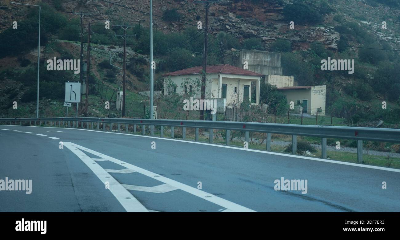 Curva autostradale asfalto bagnato sulla rotta VOAK, Creta. Presenta chiare linee di demarcazione bianche, cartelli stradali e una casa tradizionale contro una montagna rocciosa Foto Stock