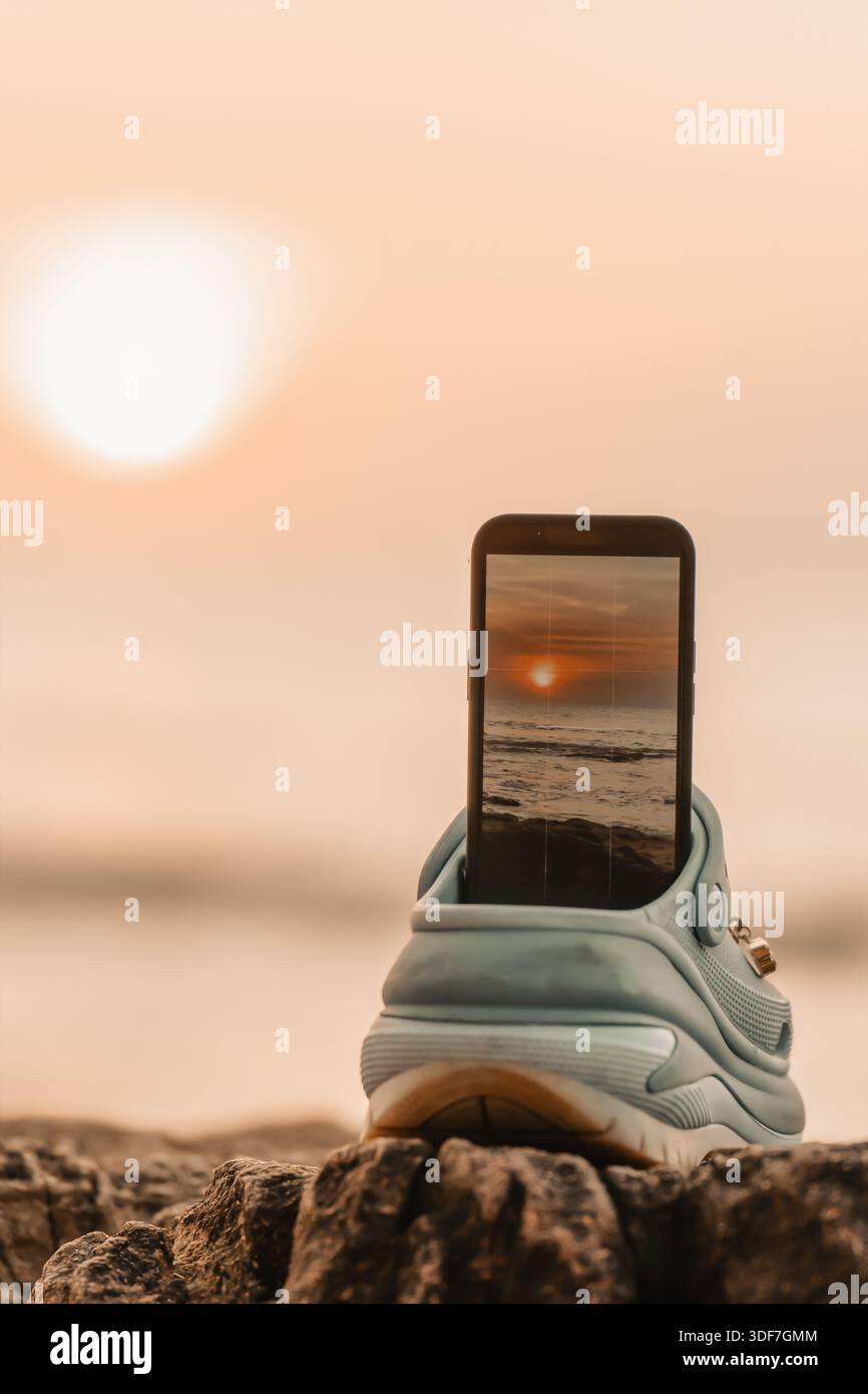 Uno smartphone mostra un'immagine del tramonto mentre si riposa su una sneaker posizionata su un terreno roccioso di una spiaggia in Thailandia durante la luce serale. Si possono vedere le onde Foto Stock