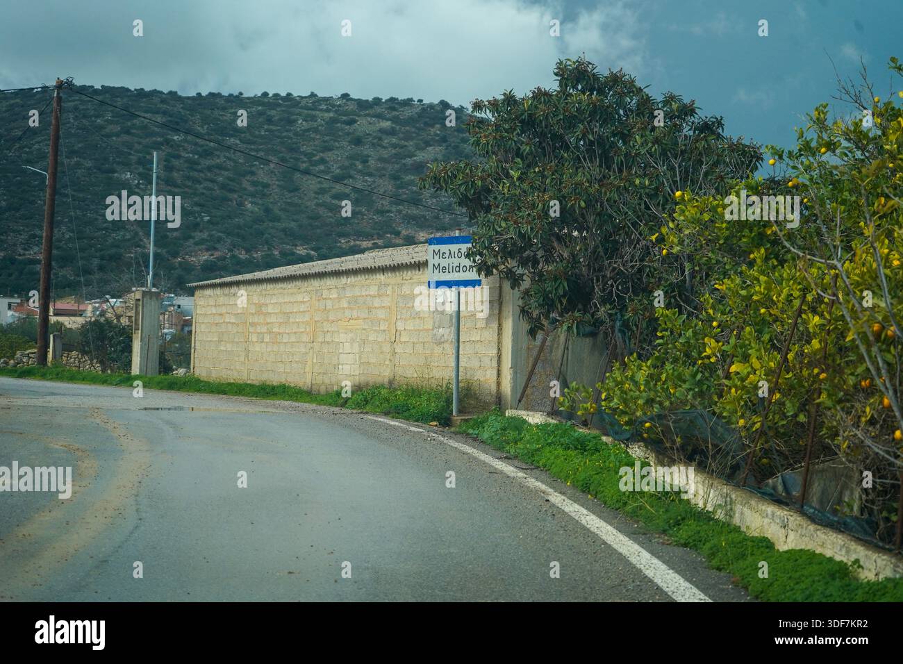 Un cartello stradale che segna l'ingresso al paese di Melidoni, Creta. Circondato da una lussureggiante vegetazione verde, alberi di agrumi e da un tradizionale muro di pietra Foto Stock